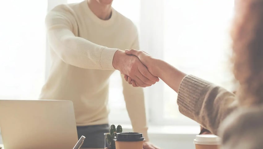 Man and woman shaking hands near a table, representing partnership and hiring - ‘Why,’ ‘When,’ and ‘How’ of Hiring Workers for Your Insurance Agency